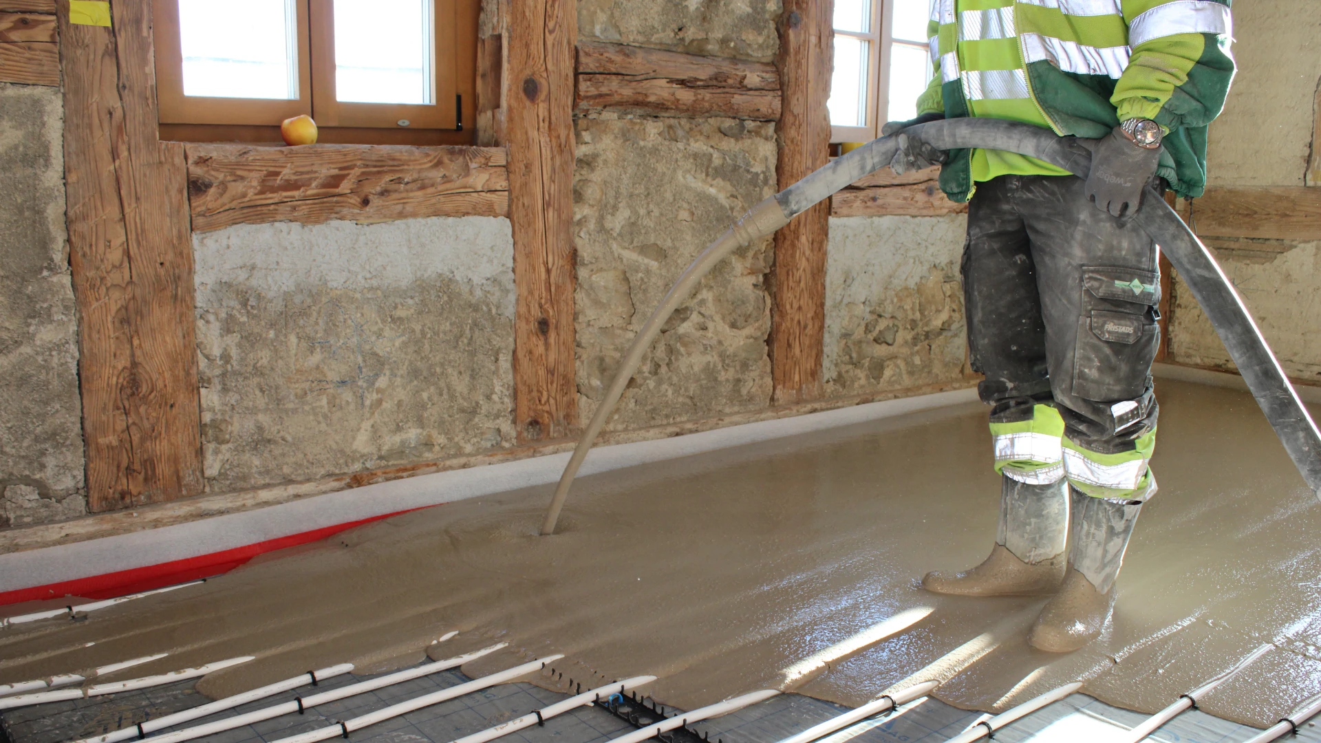 Eine Person in Arbeitskleidung verteilt fliessenden Bodenbelag über einer verlegten Bodenheizung in einem Raum mit sichtbar alter Holzwand. Der frische Belag verteilt sich gleichmässig über die Oberfläche.